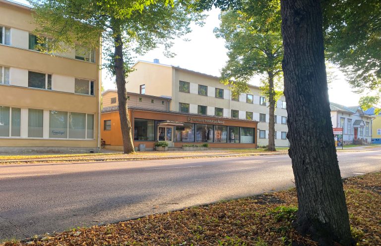 I förgrunden en höstlövsbeströdd gräsmatta och stammen hos en lönn. Bortanför, en väg, på andra sidan vägen syns NIPÅs verksamhetshus med orange fasad. Huset är omgivet av bostadshus och grönskande träd.